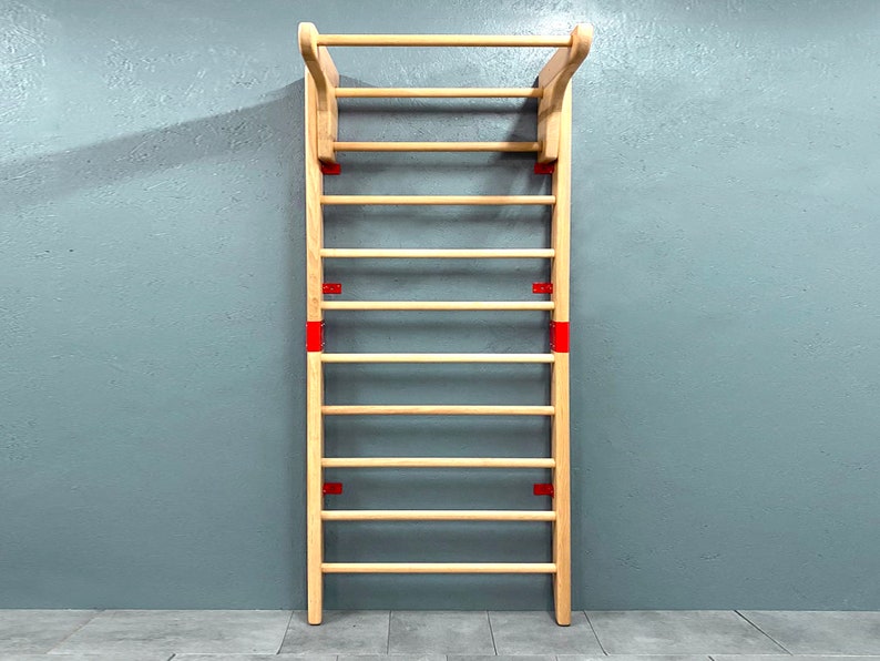 May include: A wooden wall-mounted stall bar with a horizontal top bar and multiple rungs. The wooden frame is light-colored, with red metal brackets securing the rungs. The stall bar is designed for exercise and stretching.