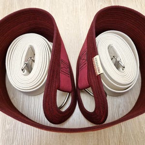 Puede incluir: Dos correas de yoga color burdeos con cinta de algodón blanca y hebillas metálicas plateadas. Las correas están enrolladas y el nombre de la marca "ProYoga" está impreso en una correa.