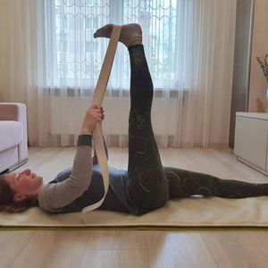 May include: A woman is lying on a white yoga mat with her right leg raised and held in place with a beige yoga strap. She is wearing black and green leggings and a gray shirt.