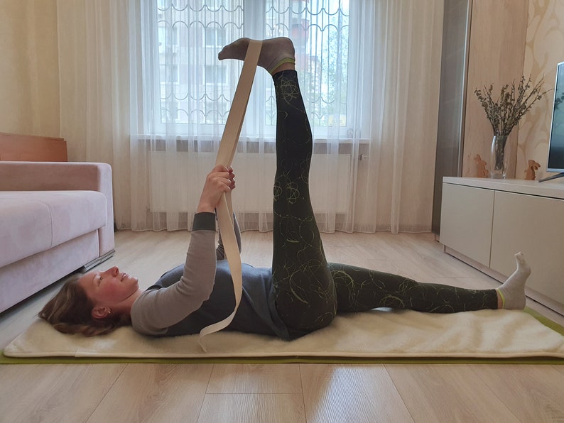 May include: A woman is lying on a white yoga mat with her right leg raised and held in place with a beige yoga strap. She is wearing black and green leggings and a gray shirt.