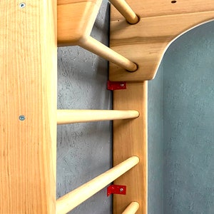 May include: Wooden climbing frame with horizontal dowels and vertical supports. The frame is made of light-colored wood, with red metal brackets securing the dowels. The frame is mounted on a textured gray wall.