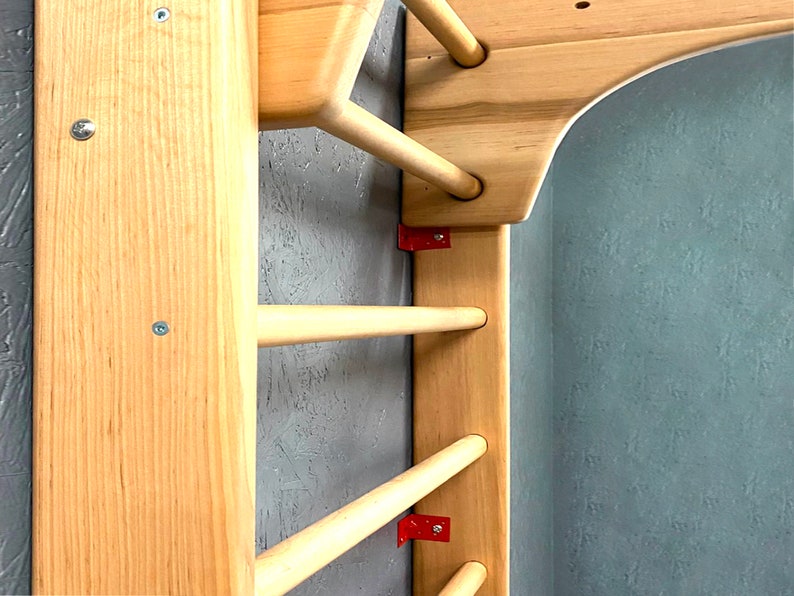 May include: Wooden climbing frame with horizontal dowels and vertical supports. The frame is made of light-colored wood, with red metal brackets securing the dowels. The frame is mounted on a textured gray wall.