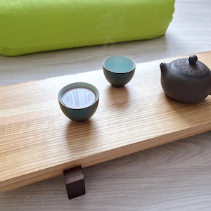 May include: A wooden serving tray with two small teal-colored teacups and a black teapot. The tray has two dark wood legs and is sitting on a light gray surface.