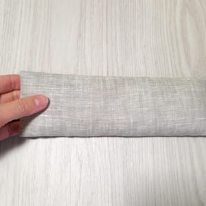 May include: A rectangular, light grey linen eye pillow. The pillow is held by a hand and has a small tag that reads "PROYOGIV". The pillow is approximately 20 cm long.