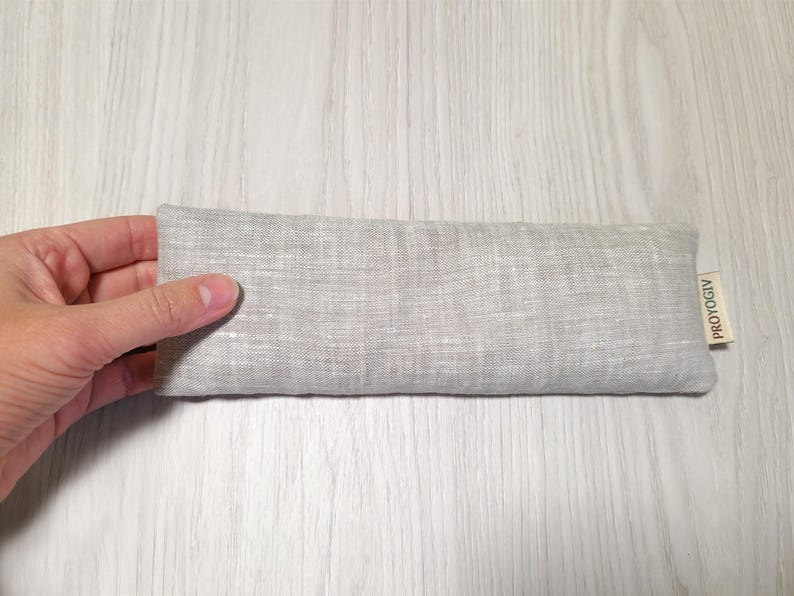 May include: A rectangular, light grey linen eye pillow. The pillow is held by a hand and has a small tag that reads "PROYOGIV". The pillow is approximately 20 cm long.