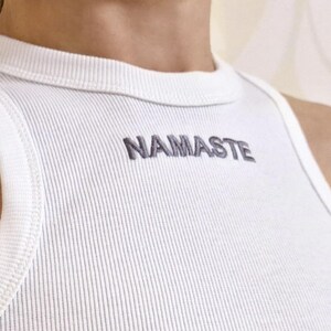 K&ouml;nnte beinhalten: Wei&szlig;es geripptes Tanktop mit dem Wort "NAMASTE" in Grau auf der Vorderseite gestickt.