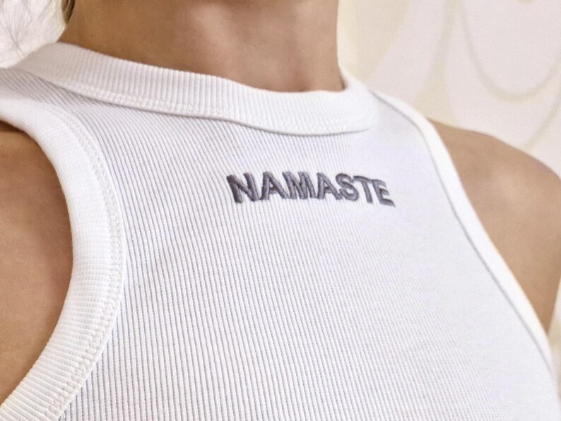 K&ouml;nnte beinhalten: Wei&szlig;es geripptes Tanktop mit dem Wort "NAMASTE" in Grau auf der Vorderseite gestickt.