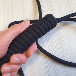 May include: A black rope with a textured handle is held in a hand. The handle is tightly wrapped with black cord, creating a ridged surface for grip. The rope extends out of frame.