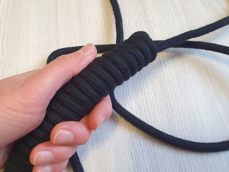 May include: A black rope with a textured handle is held in a hand. The handle is tightly wrapped with black cord, creating a ridged surface for grip. The rope extends out of frame.