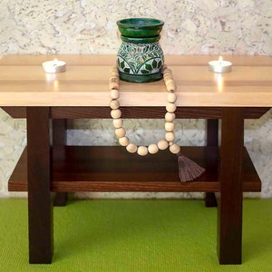 Può includere: Un piccolo tavolo di legno con un piano in legno chiaro e gambe e ripiano in legno scuro. Un bruciatore di incenso in ceramica verde con un motivo floreale si trova sullo scaffale superiore, insieme a due candele bianche e una collana di perline di legno.