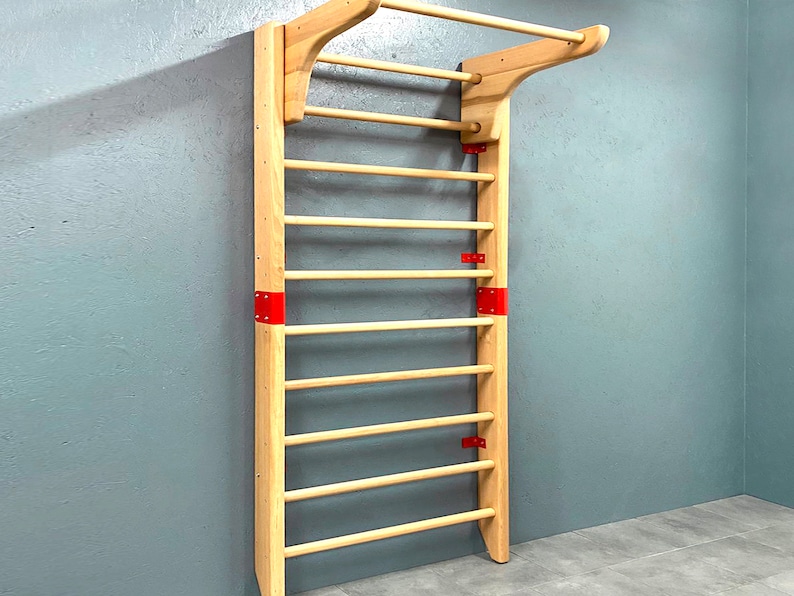 May include: A wooden stall bar with a pull-up bar at the top. The frame is light-colored wood, with horizontal dowels for climbing. Red metal brackets reinforce the frame. The stall bar is mounted on a gray wall.