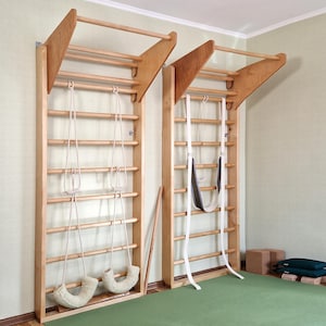 Op de afbeelding: Twee houten Zweedse ladders aan de muur met bevestigde touwen en schommels. De ladders zijn gemaakt van lichtgekleurd hout en zijn gemonteerd op een lichtgroene muur. De vloer is bedekt met een groene mat. Extra trainingsapparatuur is zichtbaar.
