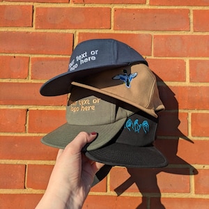 Könnte beinhalten: Ein Stapel von Five-Panel-Caps in verschiedenen Farben, darunter Marineblau, Beige, Olivgrün und Schwarz. Einige Caps haben gestickte Designs, darunter eine Ente und Gebärdensprachsymbole. Der Text "Your text or logo here" ist auf einigen Caps gestickt.