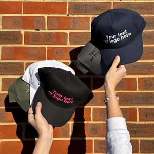 Puede incluir: Tres gorras de béisbol en blanco con correas ajustables. Las gorras son negras, azul marino y blancas. Las gorras tienen el texto "Your text or logo here" bordado en blanco y rosa.