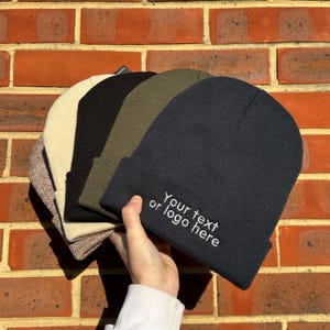 May include: Four knit beanies in different colours, including beige, black, olive green, and navy blue. The navy beanie has white embroidery that reads "Your text or logo here".
