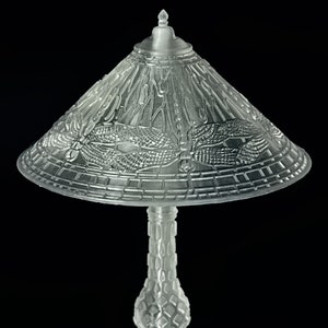 Puede incluir: Una lámpara de mesa de vidrio transparente con un diseño decorativo de libélula en la pantalla. La lámpara tiene una base redonda y un tallo alto y delgado.