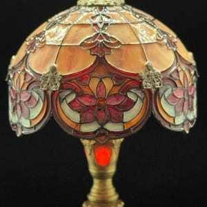 May include: A stained glass lamp with a domed shade. The shade features a geometric pattern in shades of orange, red, and pink, with floral accents. The base is a brass color. The lamp is lit with a red glow.
