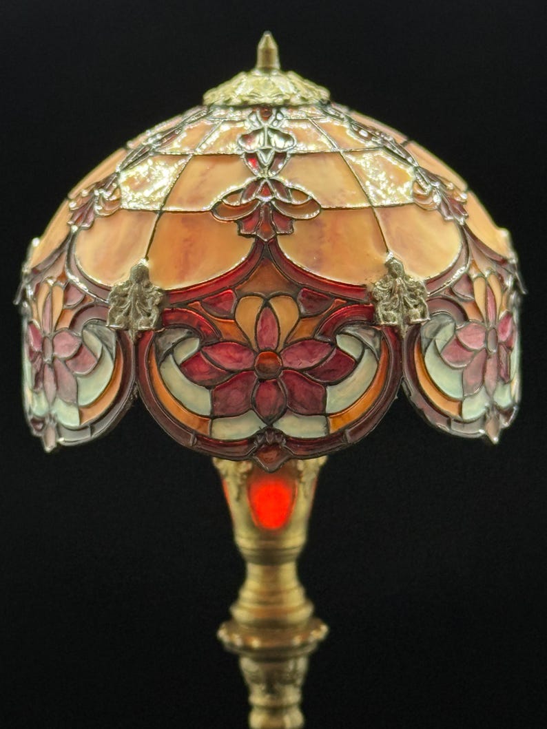 May include: A stained glass lamp with a domed shade. The shade features a geometric pattern in shades of orange, red, and pink, with floral accents. The base is a brass color. The lamp is lit with a red glow.