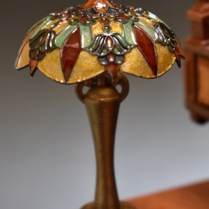 K&ouml;nnte beinhalten: Eine Miniatur-Tiffanylampe mit goldener Basis und einem bunten Schirm mit gr&uuml;nen, braunen und goldenen Akzenten. Die Lampe steht auf einer Holzoberfl&auml;che.