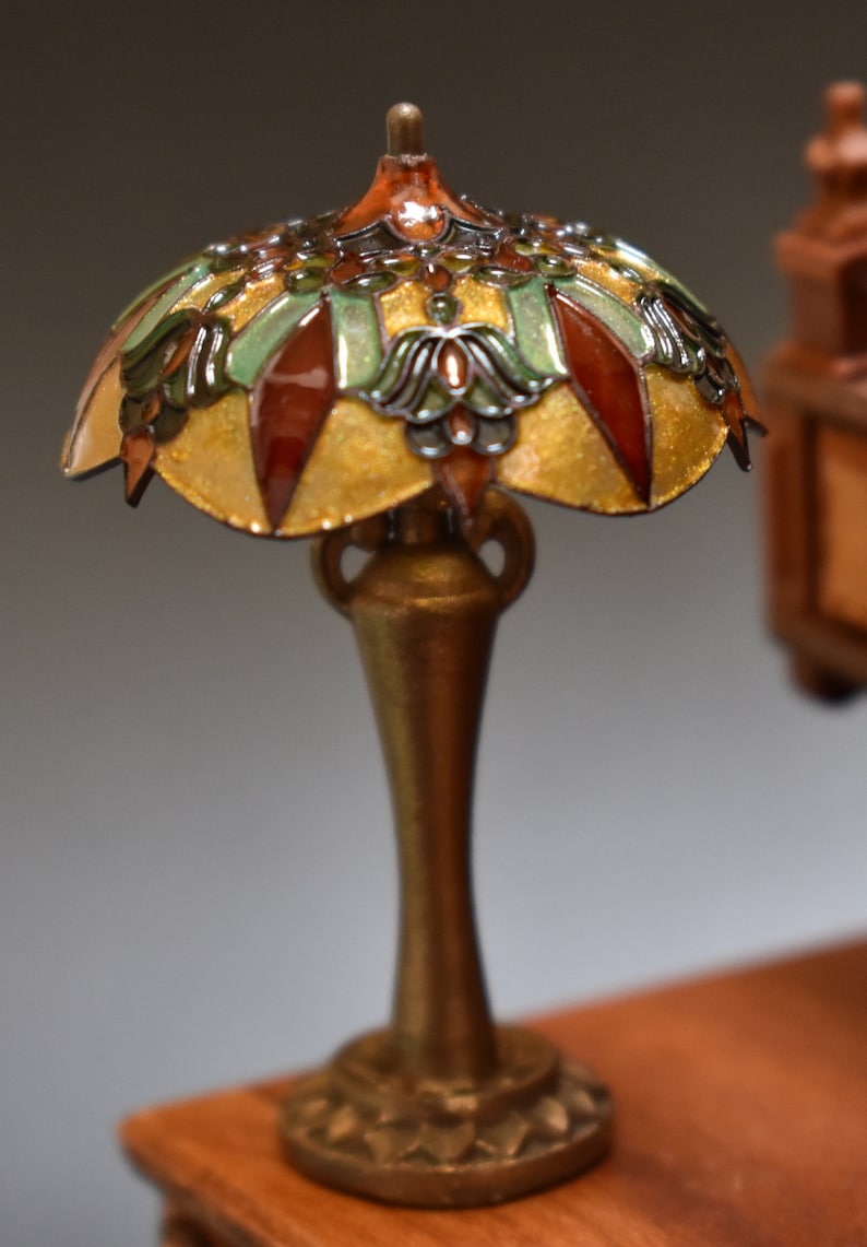 K&ouml;nnte beinhalten: Eine Miniatur-Tiffanylampe mit goldener Basis und einem bunten Schirm mit gr&uuml;nen, braunen und goldenen Akzenten. Die Lampe steht auf einer Holzoberfl&auml;che.