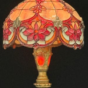 May include: A stained glass table lamp with a floral design. The lamp features a domed shade with red, orange, and cream-colored glass panels. The base is gold-colored with ornate detailing. The lamp is a decorative lighting fixture.