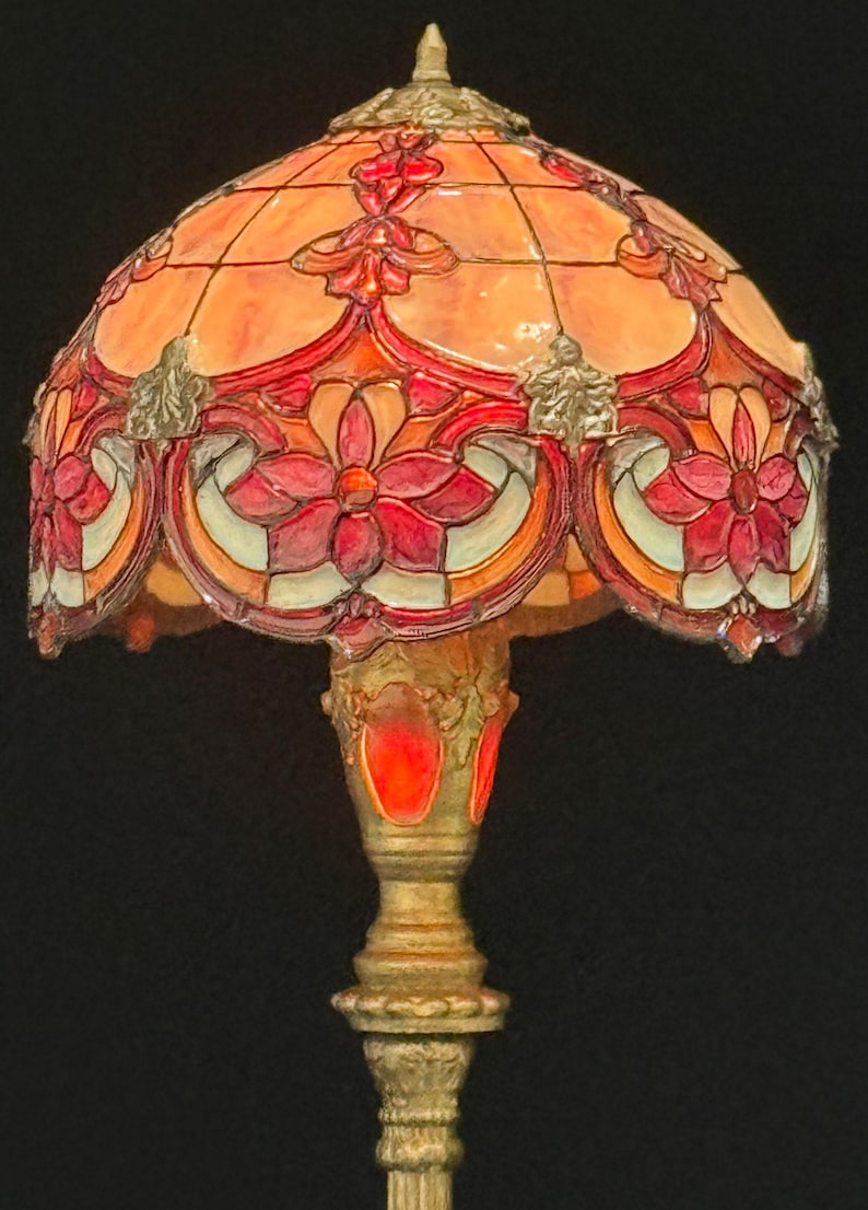 May include: A stained glass table lamp with a floral design. The lamp features a domed shade with red, orange, and cream-colored glass panels. The base is gold-colored with ornate detailing. The lamp is a decorative lighting fixture.