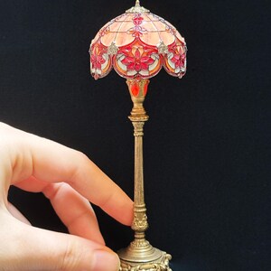 May include: Miniature Tiffany-style lamp with a stained glass shade in shades of red, orange, and pink. The lamp has a detailed brass base and stem. The lamp is held by a hand.
