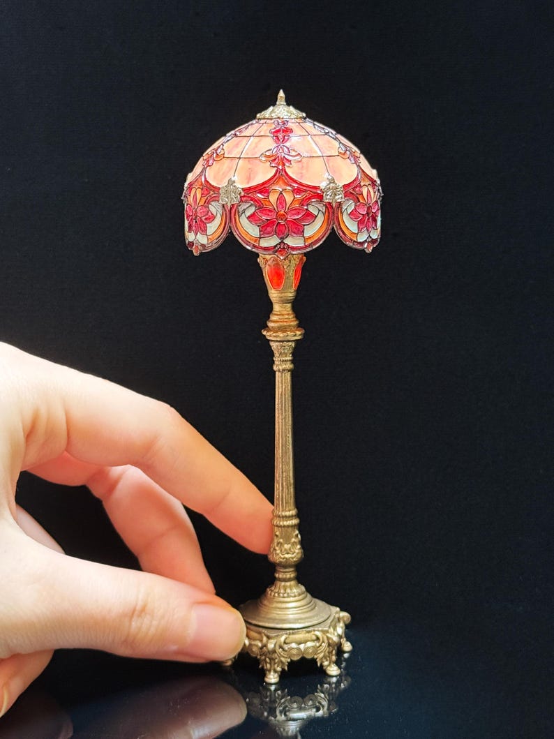 May include: Miniature Tiffany-style lamp with a stained glass shade in shades of red, orange, and pink. The lamp has a detailed brass base and stem. The lamp is held by a hand.