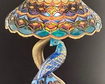 Op de afbeelding: Een miniatuur lamp van gekleurd glas met een pauwenmotief. De lampenkap is tealblauw, groen en goud van kleur met een geometrisch patroon. De voet is goudkleurig en heeft een pauw met blauwe en gouden veren.