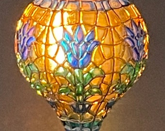 Op de afbeelding: Een hanglamp van gekleurd glas in de vorm van een traan. Het glas is een mozaïek van geel, groen, blauw en paars. De lamp is verlicht en geeft een warm licht.