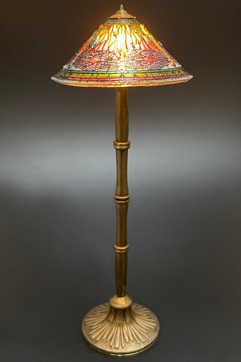 Op de afbeelding: Een gouden staande lamp met een gebrandschilderd lampenkap met een libellenmotief. De kap wordt verlicht door een warm licht.