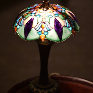 K&ouml;nnte beinhalten: Eine Buntglaslampe mit einem gr&uuml;nen, blauen, violetten und orangenen Design. Die Lampe steht auf einem Holztisch.