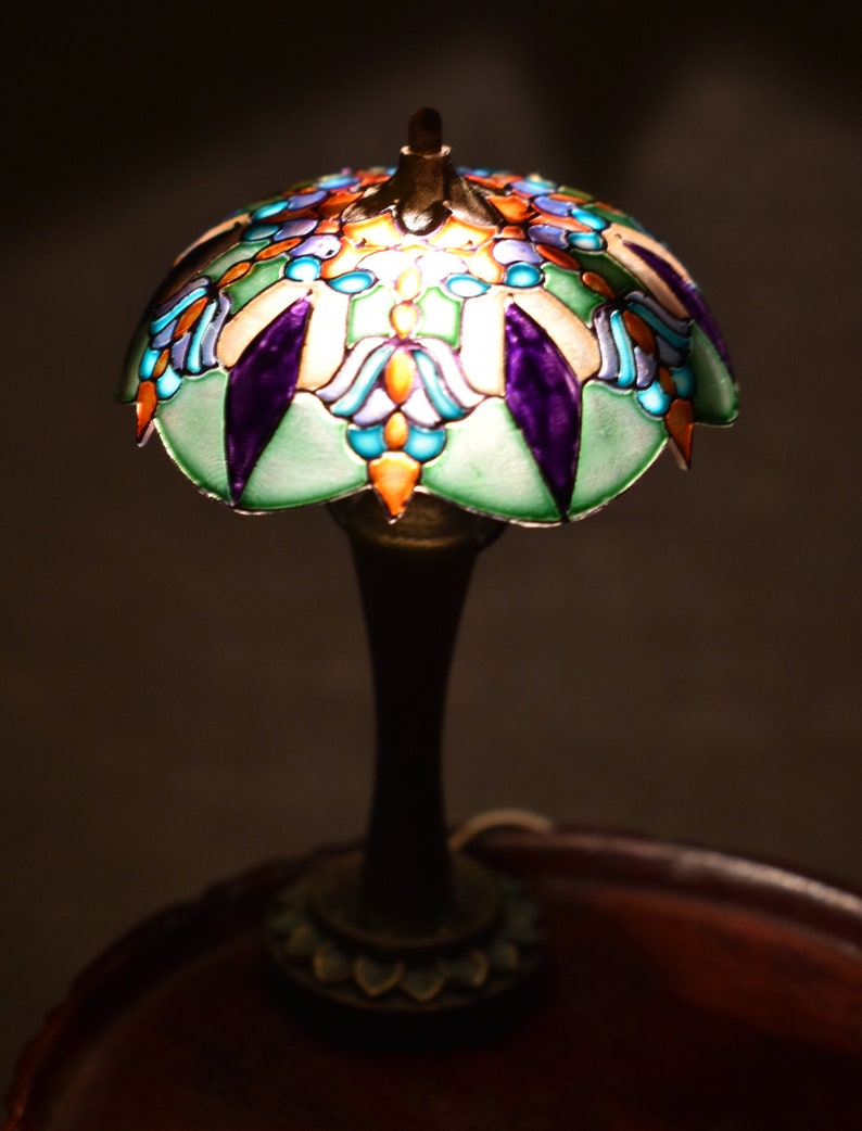 K&ouml;nnte beinhalten: Eine Buntglaslampe mit einem gr&uuml;nen, blauen, violetten und orangenen Design. Die Lampe steht auf einem Holztisch.