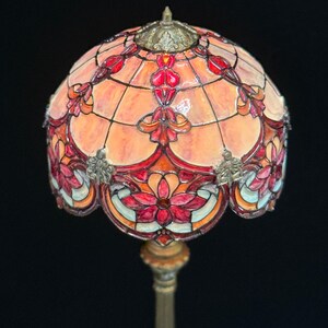 May include: A stained glass lamp with a domed shade. The shade features a floral design in shades of red, orange, and pink, with a bronze-colored metal base. The lamp is a decorative lighting fixture.