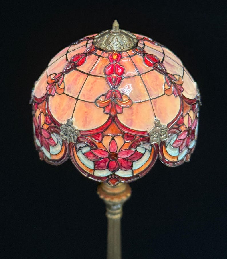 May include: A stained glass lamp with a domed shade. The shade features a floral design in shades of red, orange, and pink, with a bronze-colored metal base. The lamp is a decorative lighting fixture.
