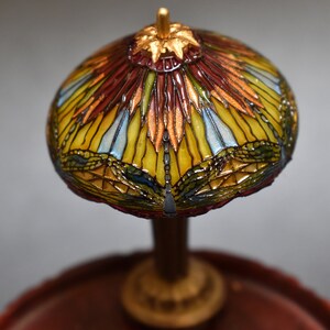 Pode incluir: Uma l&acirc;mpada de vidro Tiffany em miniatura com um design de lib&eacute;lula. O abajur &eacute; feito de pe&ccedil;as de vidro coloridas em tons de vermelho, amarelo, azul e verde. A base &eacute; de madeira marrom escura.