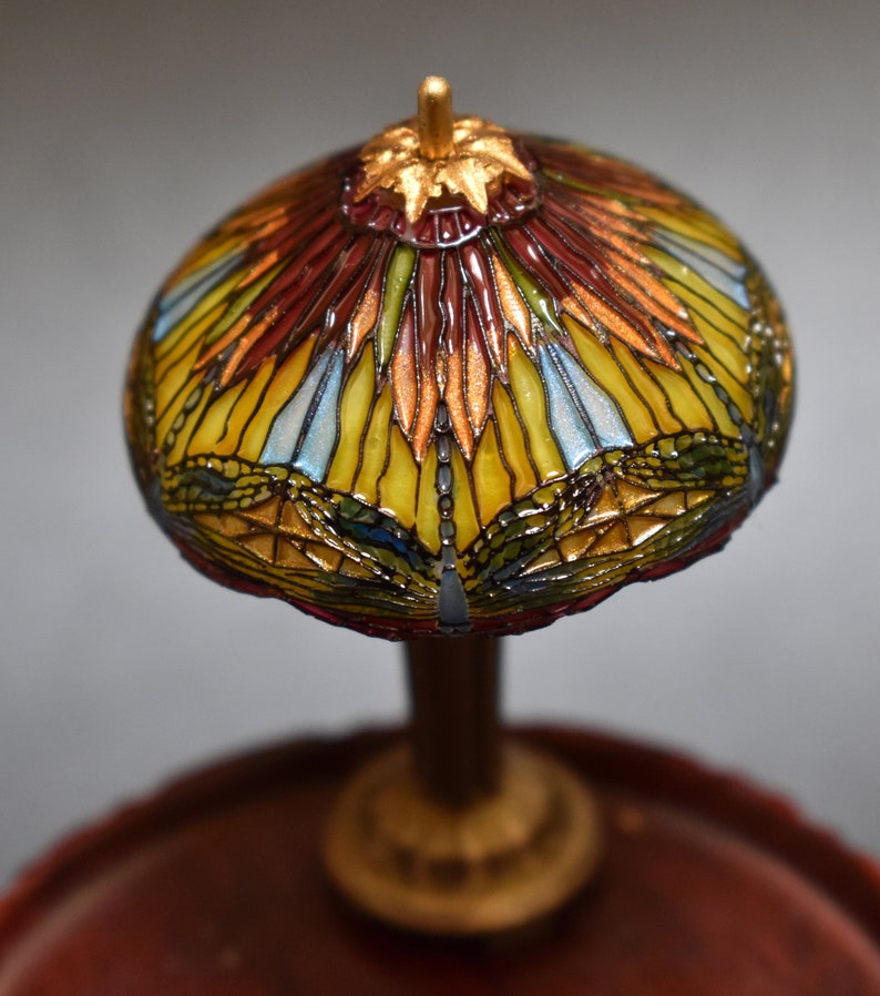 Pode incluir: Uma l&acirc;mpada de vidro Tiffany em miniatura com um design de lib&eacute;lula. O abajur &eacute; feito de pe&ccedil;as de vidro coloridas em tons de vermelho, amarelo, azul e verde. A base &eacute; de madeira marrom escura.
