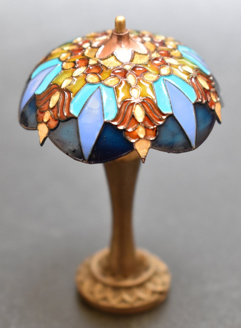 K&ouml;nnte beinhalten: Eine Miniatur-Lampe aus Buntglas mit einem blauen, t&uuml;rkisfarbenen, orangefarbenen und goldenen Design. Die Lampe hat einen goldenen Sockel.