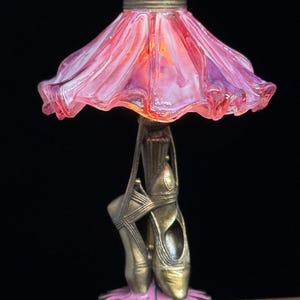 May include: A decorative lamp featuring a pink and purple glass shade and a base shaped like ballet pointe shoes. The lamp has a bronze finish and a pink floral base, creating a whimsical and elegant design.