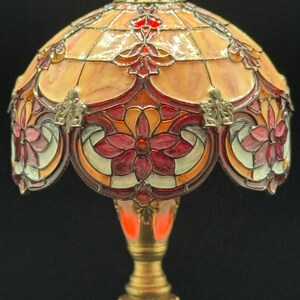 May include: A stained glass lamp with a floral design. The lamp features a domed shade with a warm color palette of orange, red, and cream. The base is a golden color, and the lamp is lit from within.