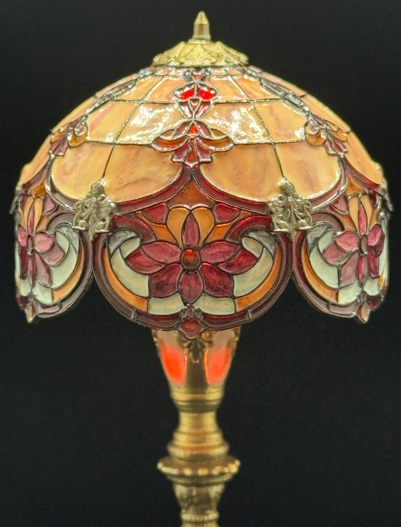 May include: A stained glass lamp with a floral design. The lamp features a domed shade with a warm color palette of orange, red, and cream. The base is a golden color, and the lamp is lit from within.