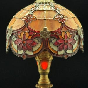 May include: A stained glass table lamp with a domed shade. The shade features a floral design in shades of orange, red, and white. The base is gold-colored with intricate detailing. The lamp is illuminated with a warm, red glow.