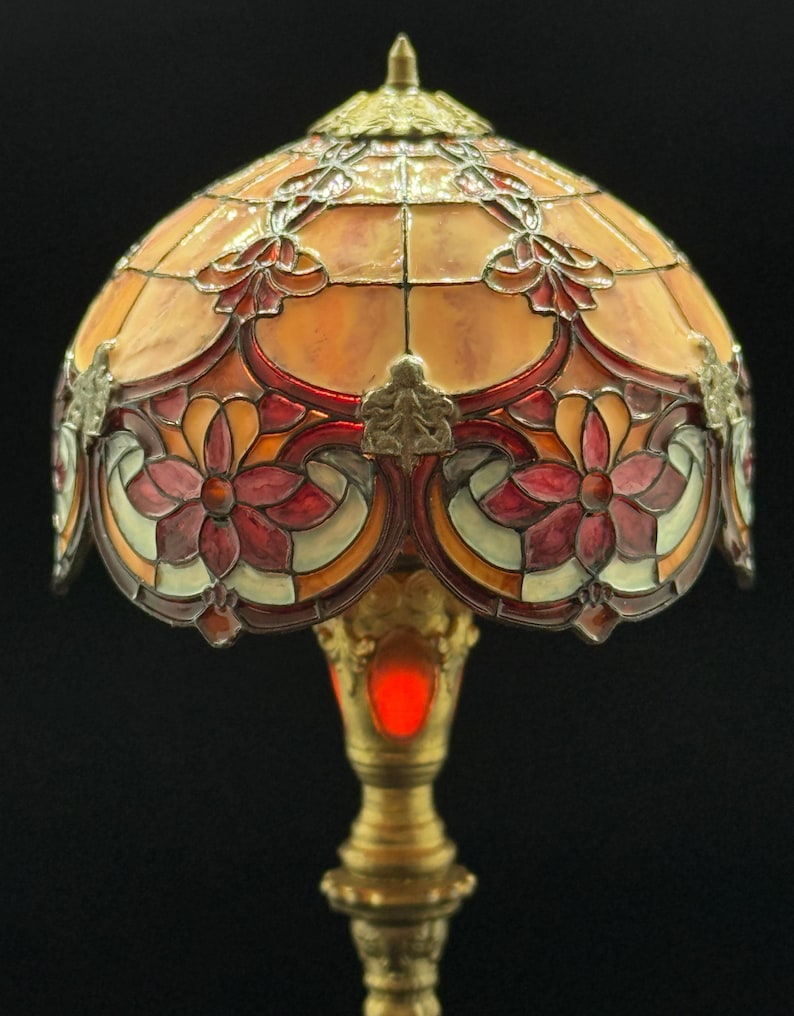May include: A stained glass table lamp with a domed shade. The shade features a floral design in shades of orange, red, and white. The base is gold-colored with intricate detailing. The lamp is illuminated with a warm, red glow.
