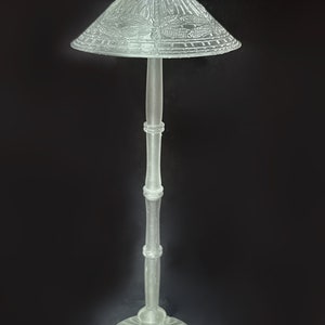Op de afbeelding: Een heldere glazen vloerlamp met een decoratieve, gepatenteerde lampenkap. De lampenkap heeft een geometrisch ontwerp en een puntige bovenkant.