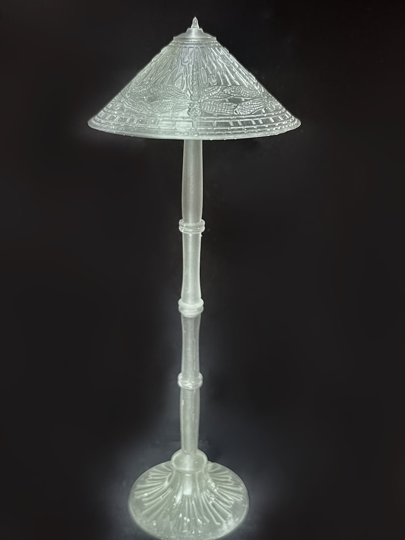 Op de afbeelding: Een heldere glazen vloerlamp met een decoratieve, gepatenteerde lampenkap. De lampenkap heeft een geometrisch ontwerp en een puntige bovenkant.