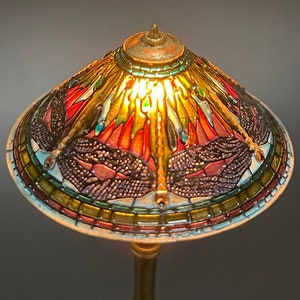 Op de afbeelding: Een lamp van gekleurd glas met een libellenmotief. De lampenkap is gemaakt van gekleurde glasstukken en heeft een gouden voet.