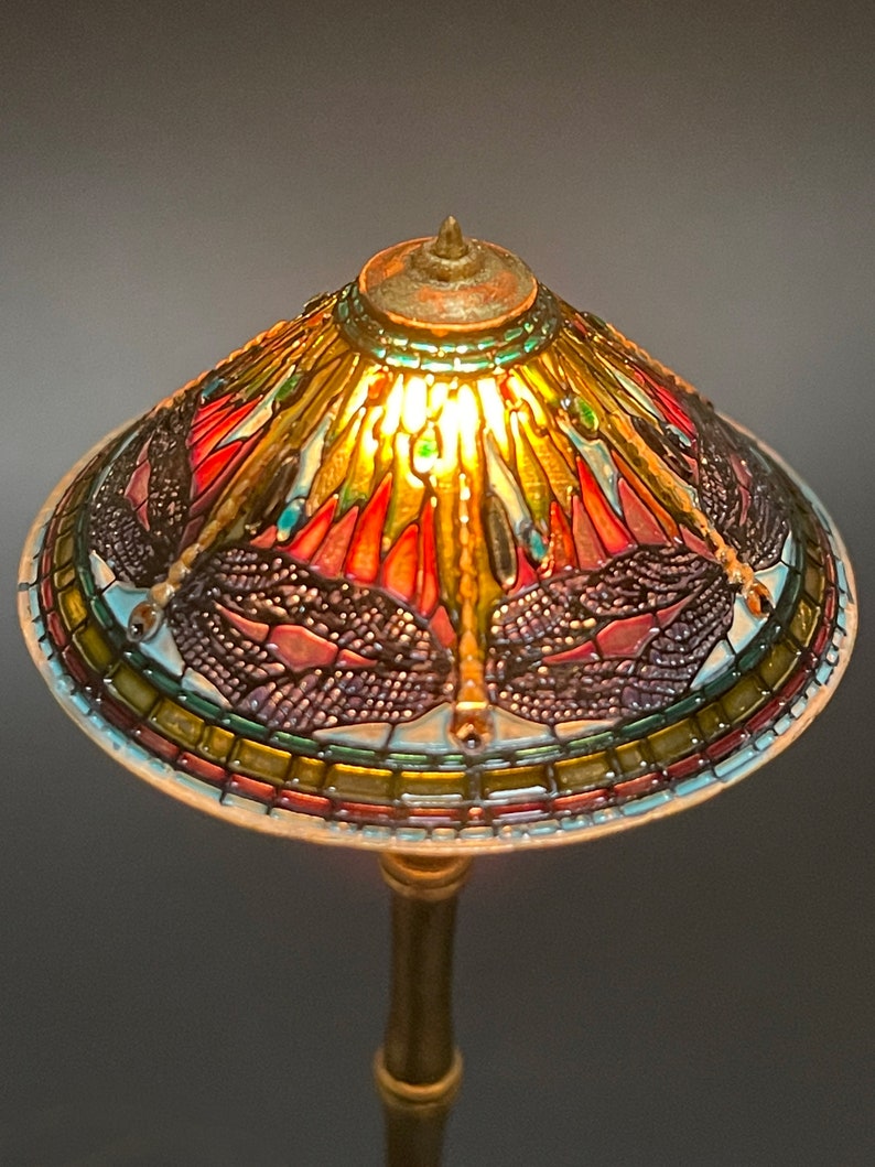 Op de afbeelding: Een lamp van gekleurd glas met een libellenmotief. De lampenkap is gemaakt van gekleurde glasstukken en heeft een gouden voet.