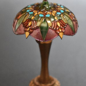 K&ouml;nnte beinhalten: Eine Miniatur-Lampe aus Buntglas mit goldener Basis. Der Lampenschirm zeigt ein florales Design in den Farben Rosa, Gr&uuml;n, Gold und T&uuml;rkis.