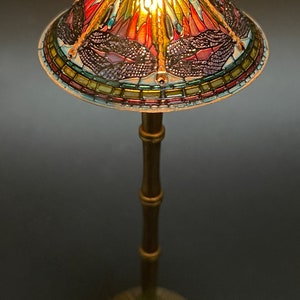 Op de afbeelding: Een tafellamp van Tiffany-glas met een libellenmotief. De lampenkap is gemaakt van gekleurde glasstukken en heeft een gouden metalen voet.