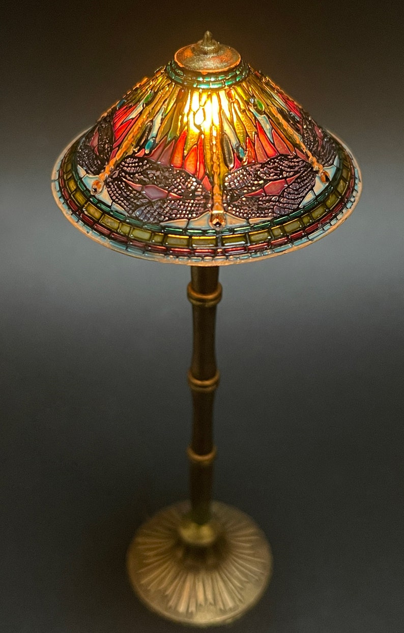 Op de afbeelding: Een tafellamp van Tiffany-glas met een libellenmotief. De lampenkap is gemaakt van gekleurde glasstukken en heeft een gouden metalen voet.